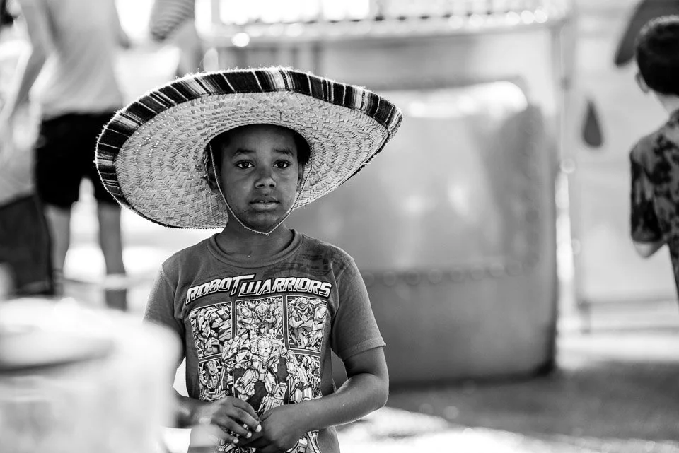 A young boy wearing a large sombrero hat and a T-shirt with comic characters stands outdoors in a busy area.