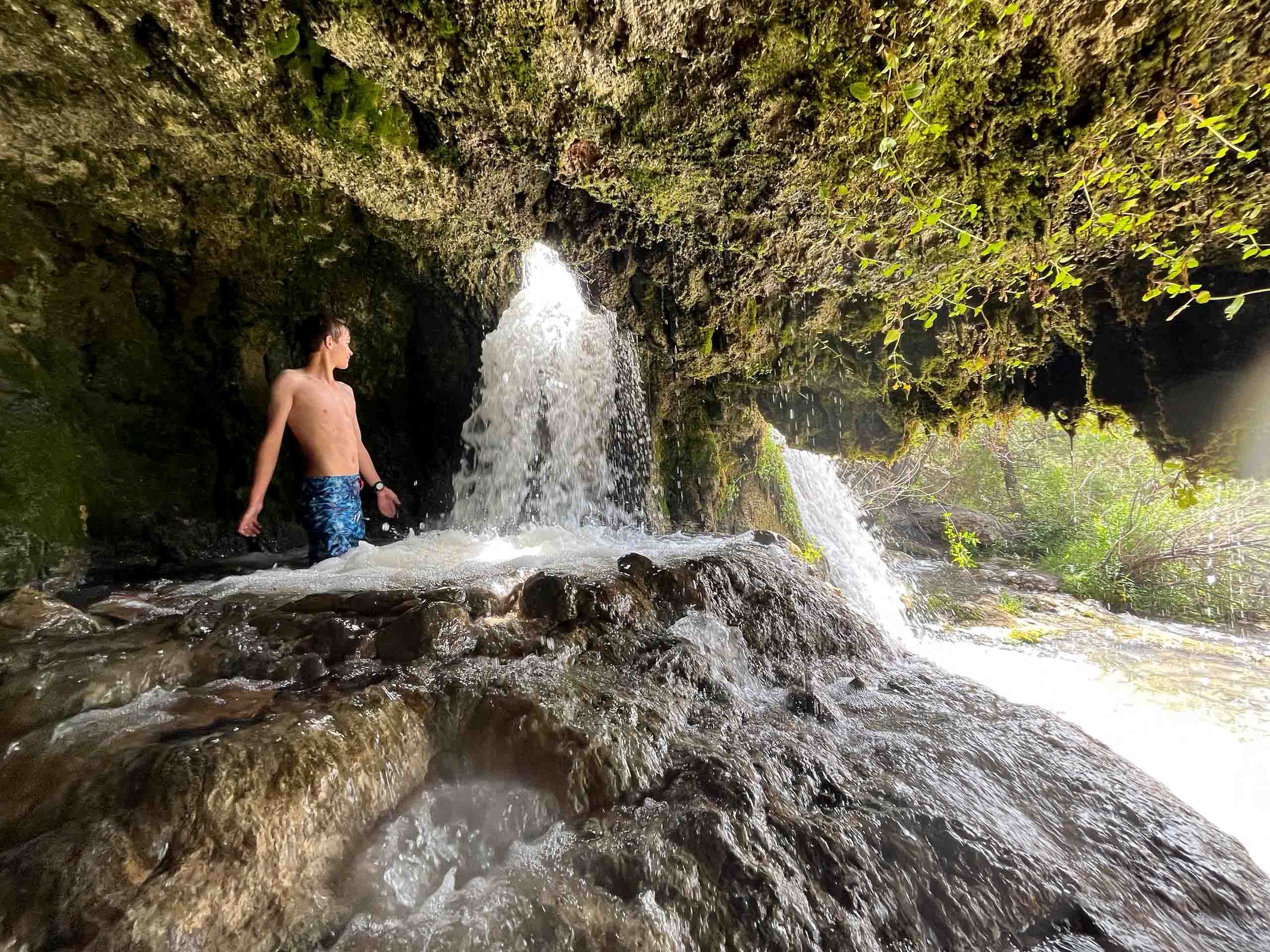 A shirtless young man in blue swim trunks standing in a small waterfall inside a rocky cave with moss-covered walls, sunlight streaming in from outside.