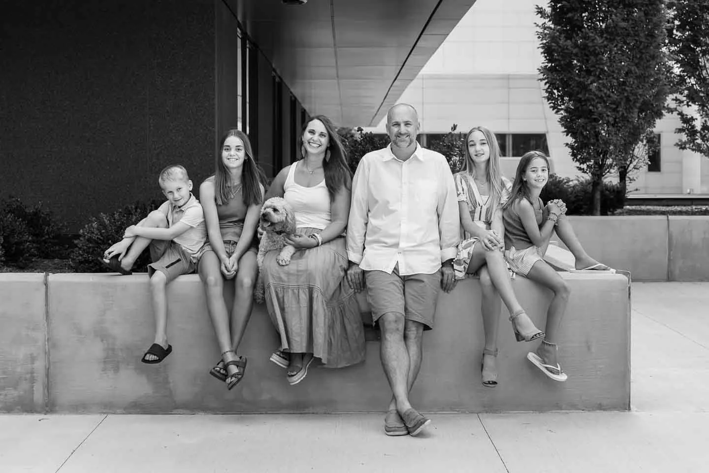 Black and white photo of a happy family of seven people and a dog sitting on a ledge outdoors in front of a modern building with trees in the background.