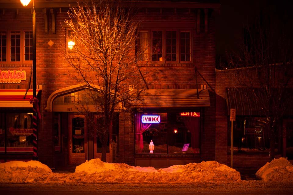 A brick building with a neon tattoo sign in the window, snow on the ground, and a leafless tree in front, illuminated by streetlights at night.