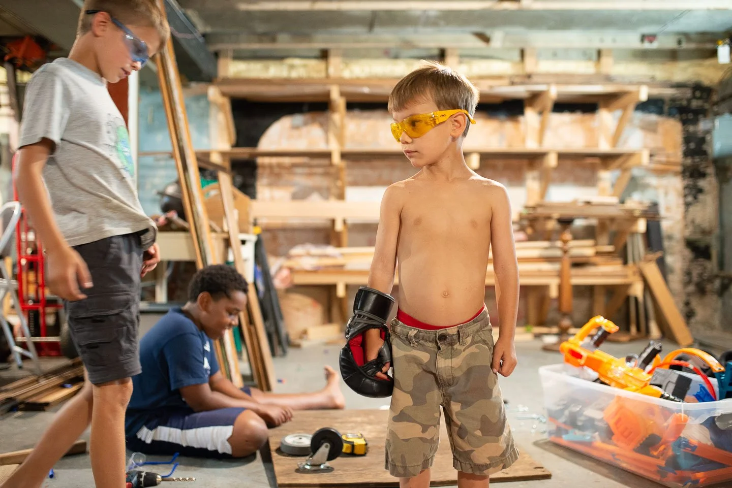 Three boys in a woodworking shop, one shirtless boy with yellow safety glasses and a glove, two other boys working with tools and wood.