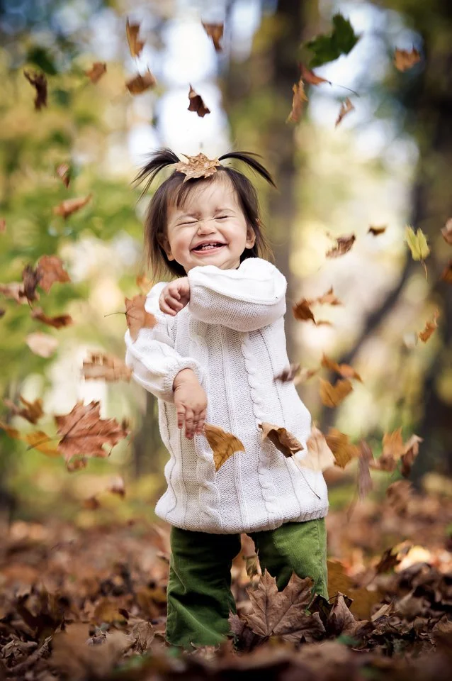 A young girl with pigtails laughing and playing in a leaf pile in a fall setting.