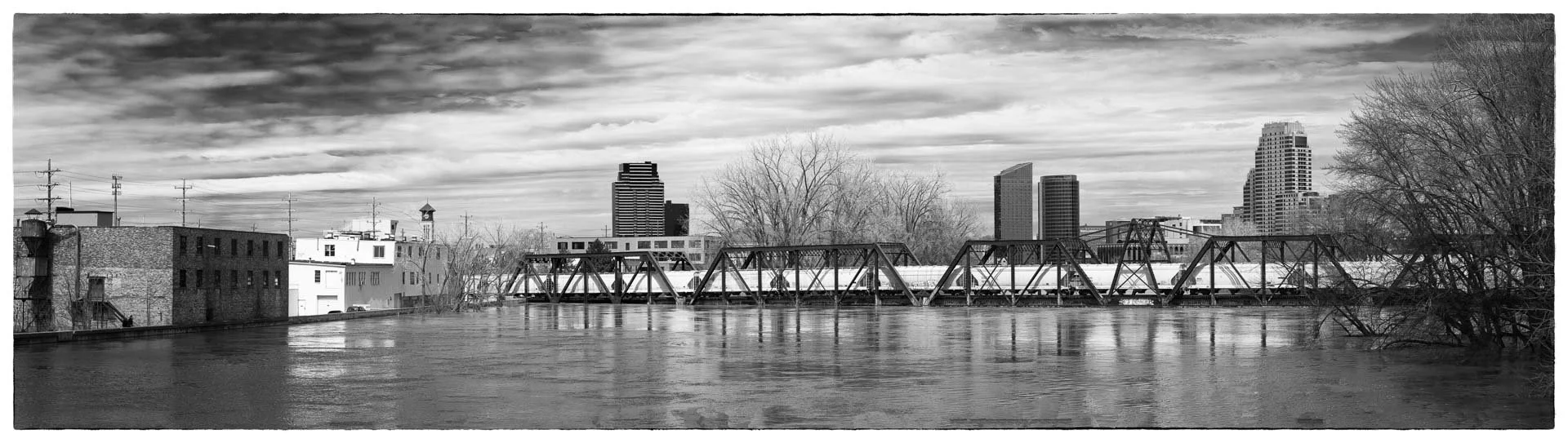 Black and white photo of a city skyline with a river in the foreground and a metal train bridge crossing the river.