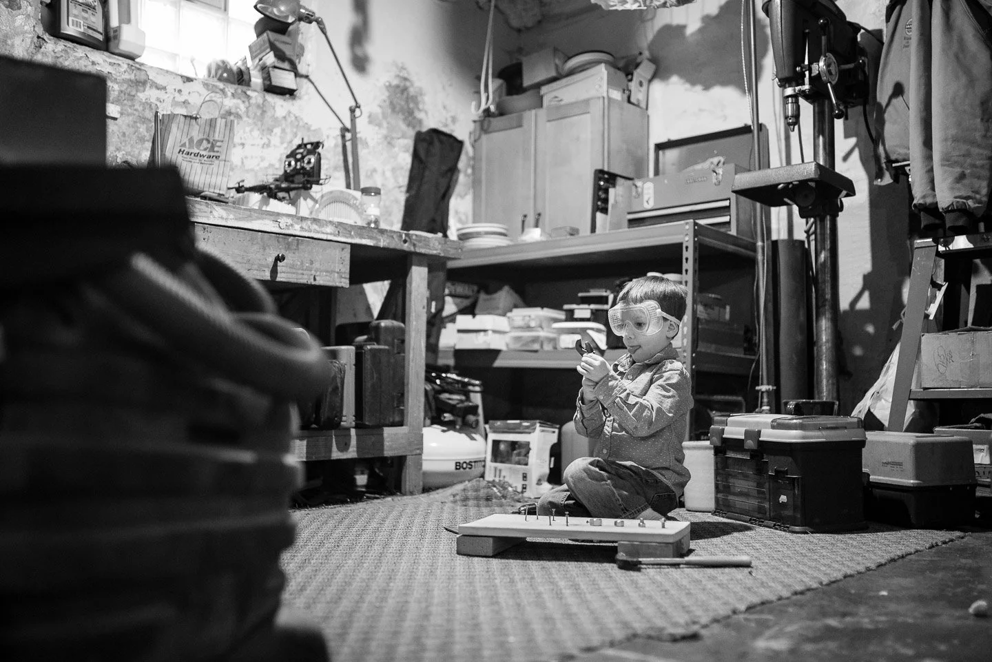 A young boy sitting on a carpeted floor in a cluttered workshop or garage, wearing safety goggles and playing with a toy car on a wooden board. There are shelves and tools visible in the background.