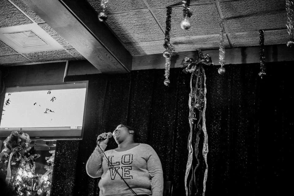 A woman singing into a microphone in a room decorated with Christmas ornaments and tinsel.