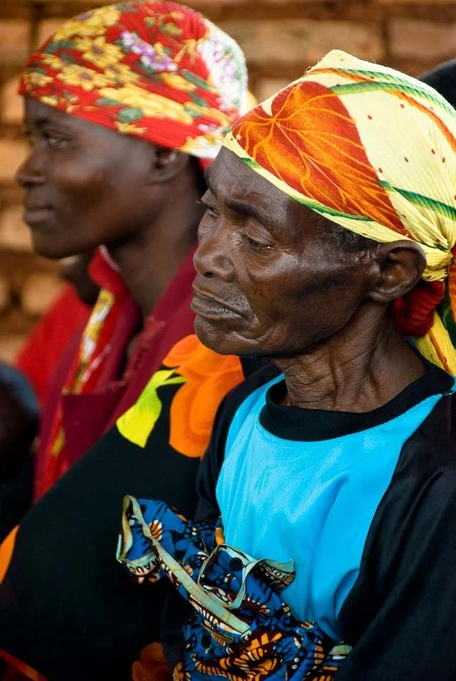 Two elderly women of African descent wearing colorful headscarves and traditional clothing, sitting side by side indoors.