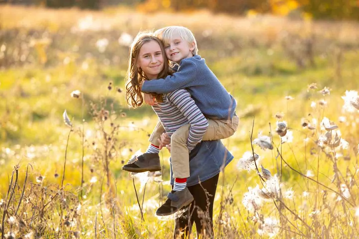 A woman carrying a young girl on her back in a field of cotton plants during autumn, with warm sunlight illuminating the scene.