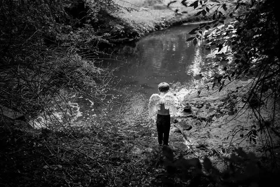 A person with angel wings standing alone on a riverbank surrounded by trees in black and white.