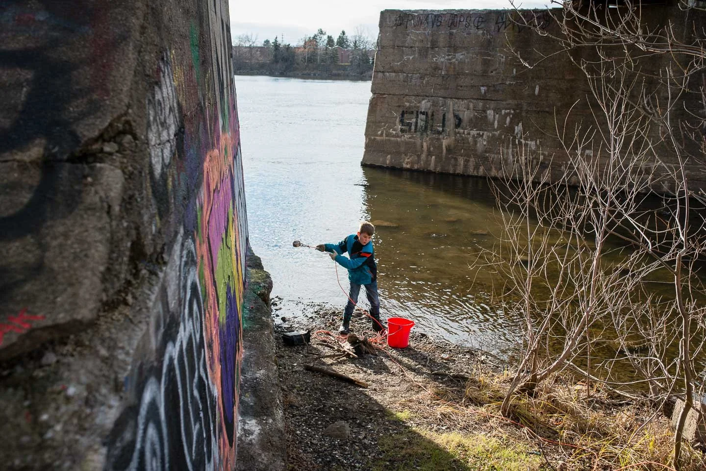A young boy wearing a blue jacket and pants is near a riverbank, holding a net with a red bucket placed on the ground nearby. In the background, there are concrete bridge supports with graffiti, and graffiti-covered brick wall on the left side of the