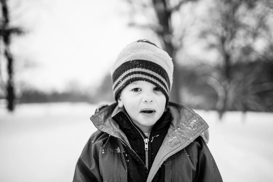 A young boy dressed in winter clothing, including a striped knit hat and a winter coat, standing outdoors in a snowy landscape with trees in the background.