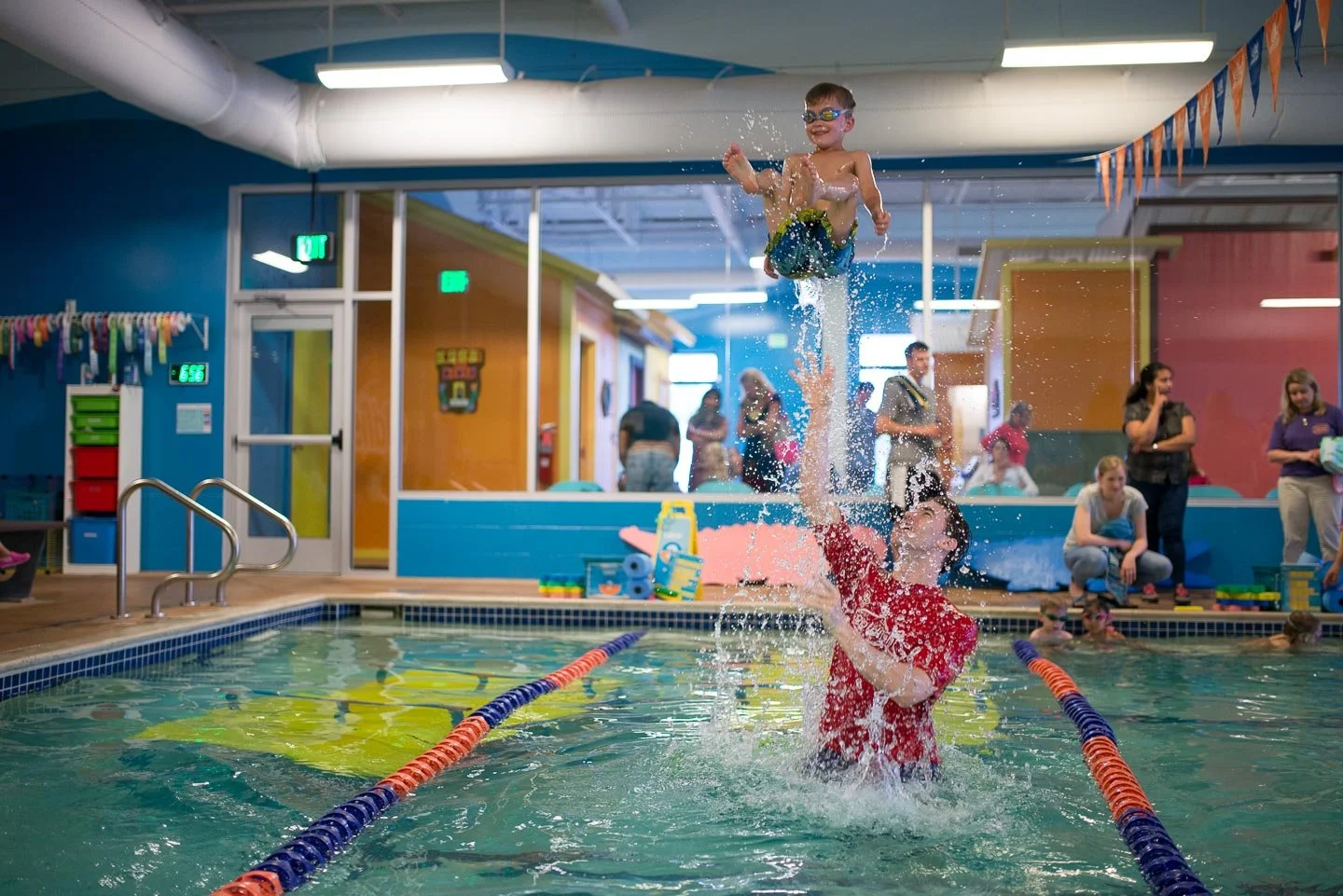 A young child wearing goggles and swim trunks being lifted out of the water by an adult woman at an indoor swimming pool, with other children and adults in the background.