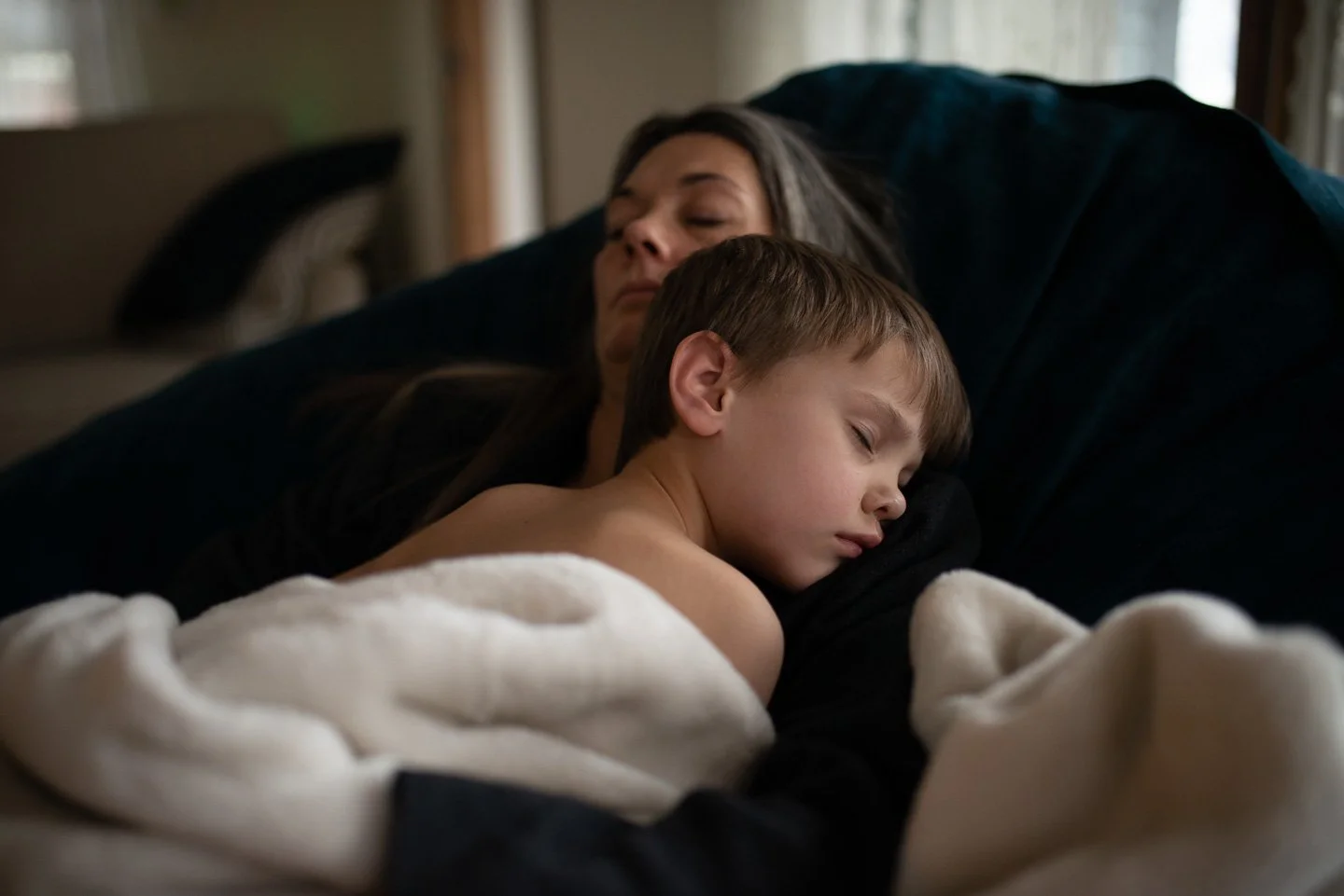 A woman and a young boy are sleeping together on a couch, snuggled under a blanket.