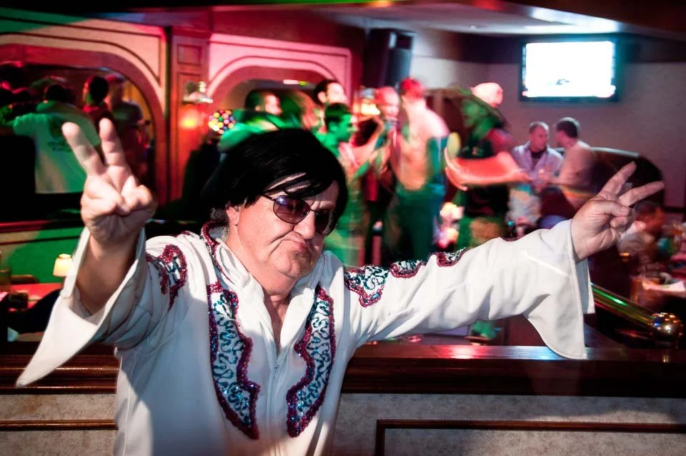 A man dressed as Elvis Presley making peace signs at a lively karaoke bar or nightclub.