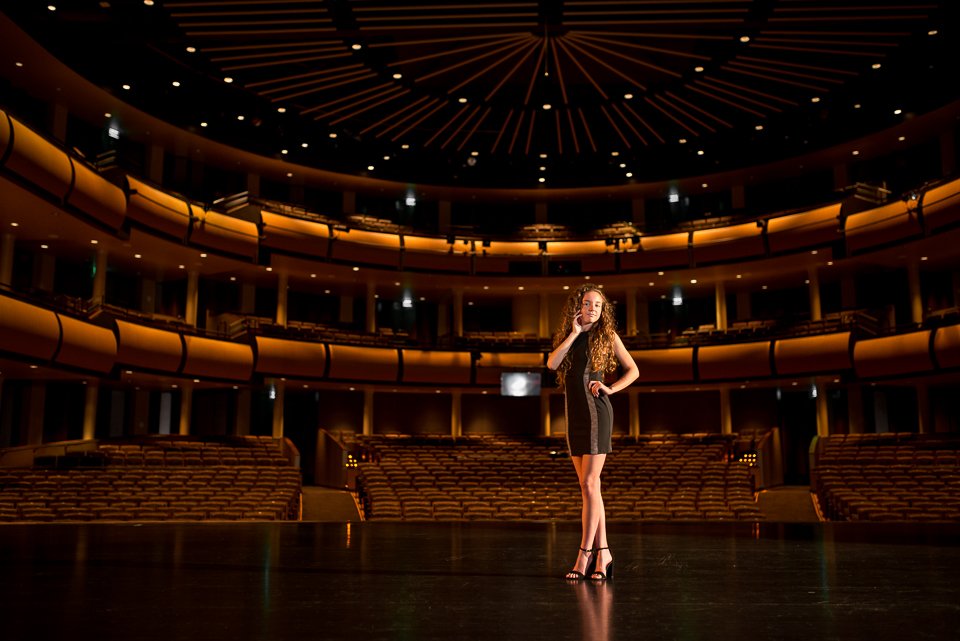 A woman in a black dress with blonde curly hair standing alone on a stage in an empty theater with multiple balconies and warm lighting.