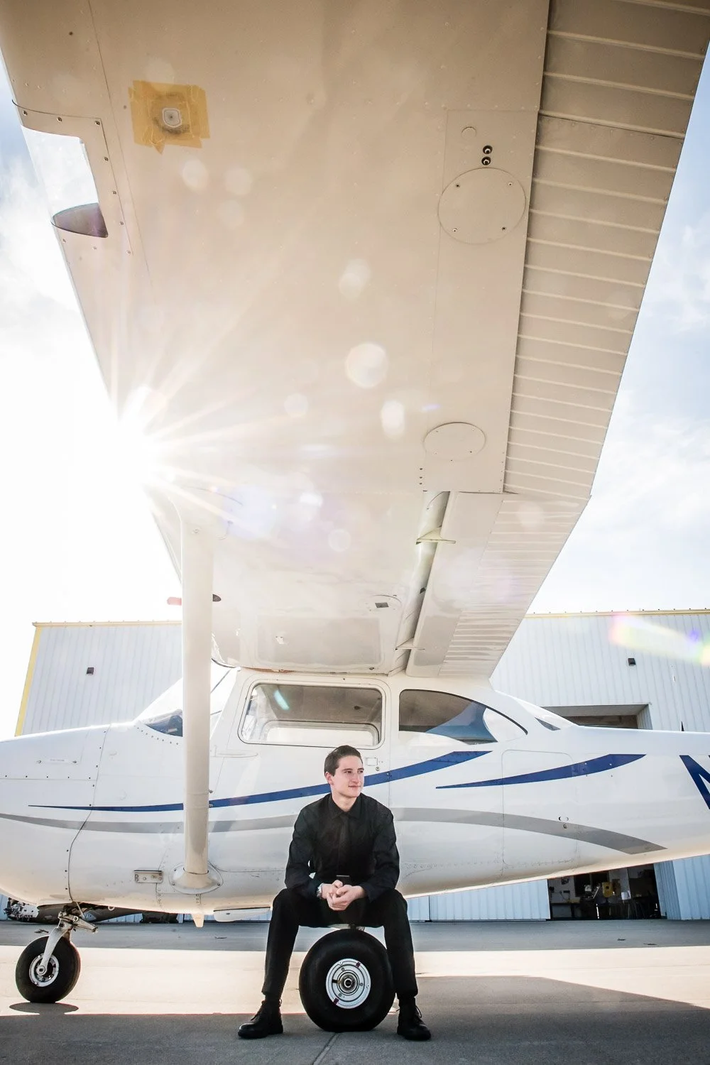 A young man in black clothing sitting on a tire in front of a small white airplane, with hangar buildings in the background and sunlight shining from the left.