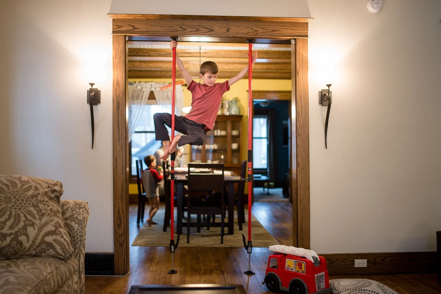 A boy hanging from a doorway frame with toy gymnastic bars in a house with two children in the background