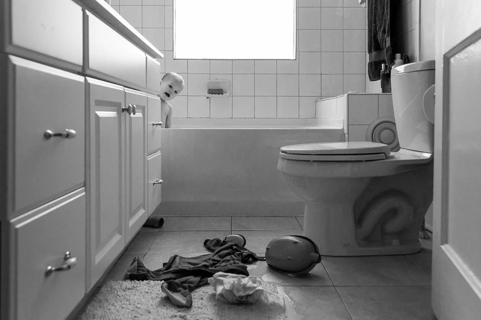 A black-and-white photo of a bathroom with a toddler peeking out from behind a cabinet, next to the bathtub. There are clothes and a toy on the floor near the toilet, and a towel hanging on a hook.