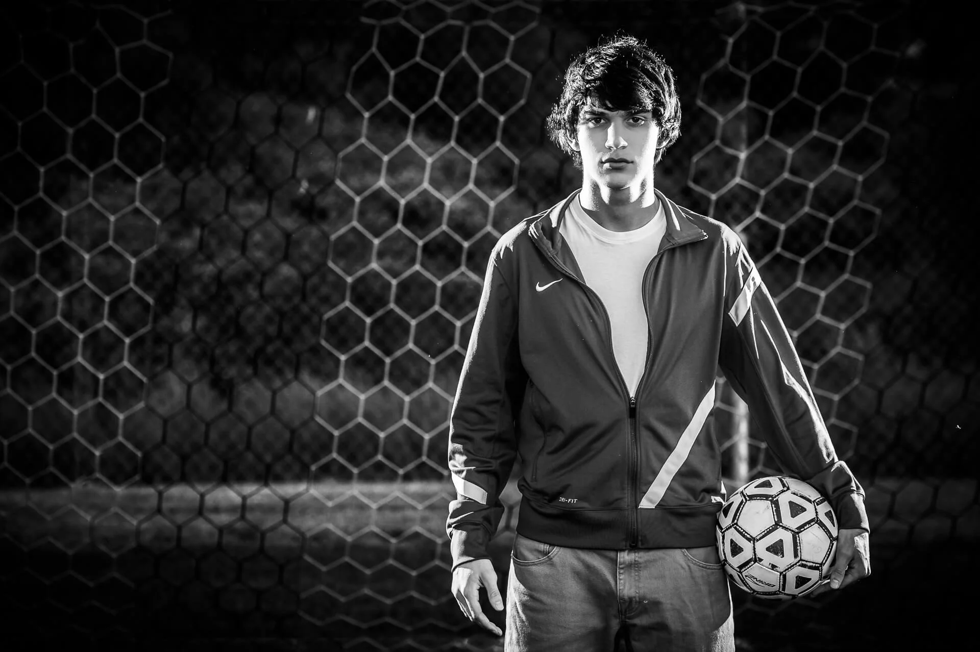 Black and white photo of a young man standing in front of a hexagonal wire fence, holding a soccer ball in his left hand, wearing a Nike sports jacket, casual jeans, and a white t-shirt.