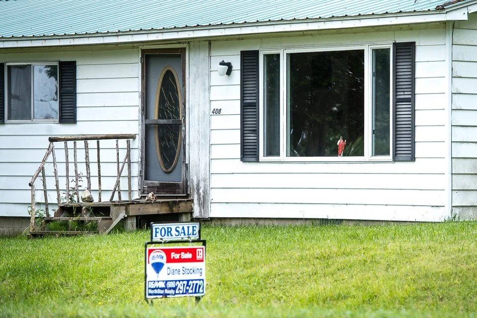 A small house with white siding, a front door with an oval glass window, a large window with black shutters, a weathered wooden front porch with steps, and a green lawn with a 'For Sale' sign in front.