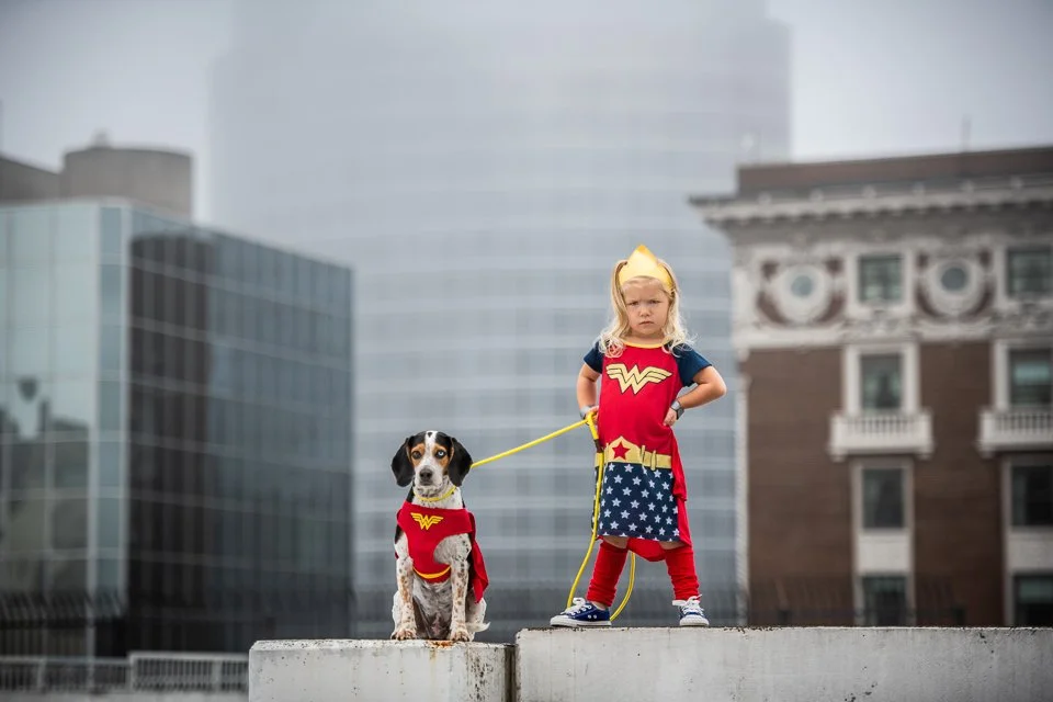 A young girl dressed as Wonder Woman standing on a concrete ledge with her dog, also dressed as Wonder Woman, in an urban setting with tall buildings in the background.