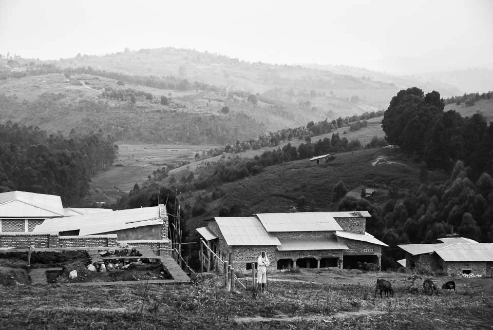 A rural scene with a woman and three goats in front of houses, with rolling hills and mountains in the background, in black and white.