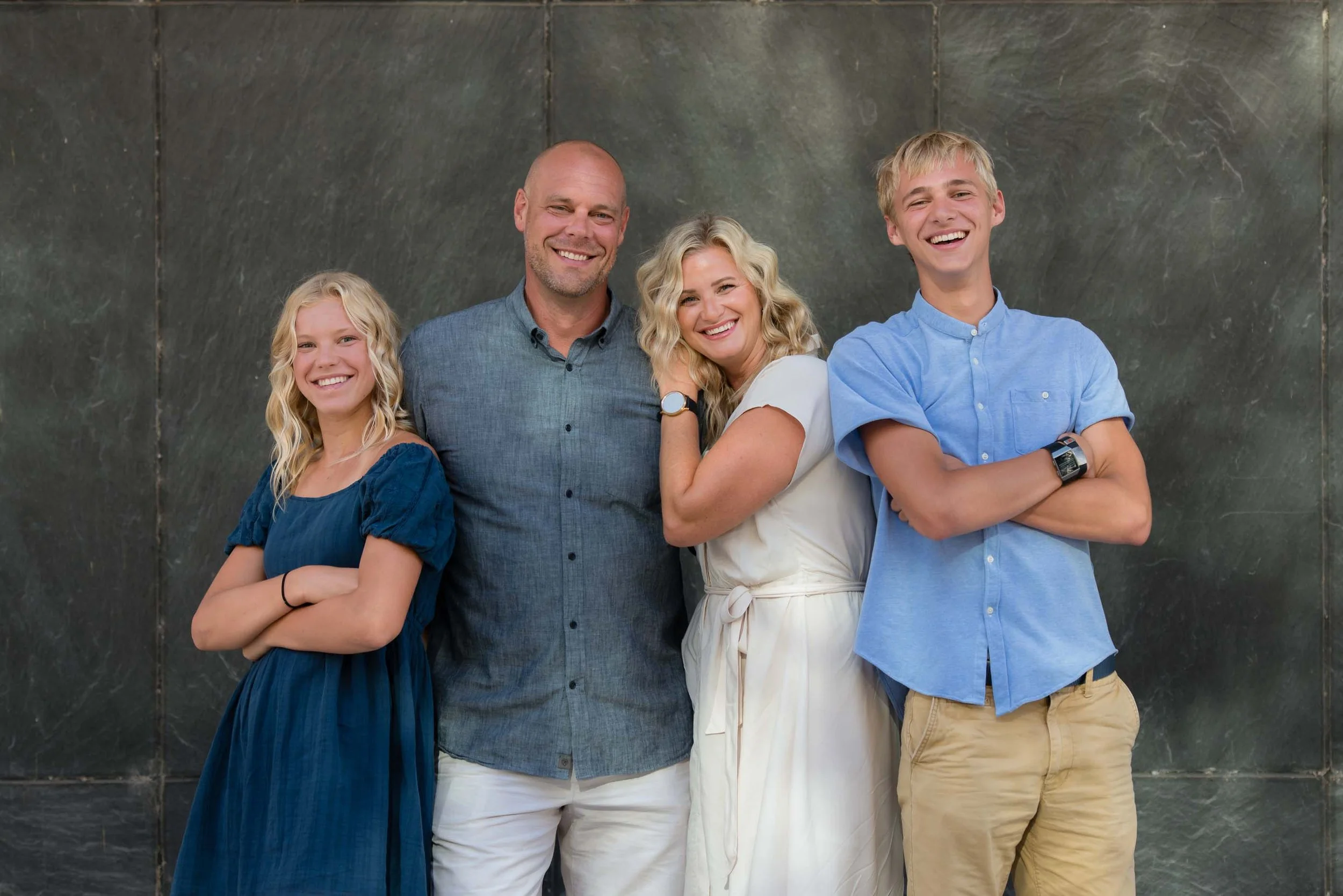 A family of four standing together, smiling, with a dark marble wall background.
