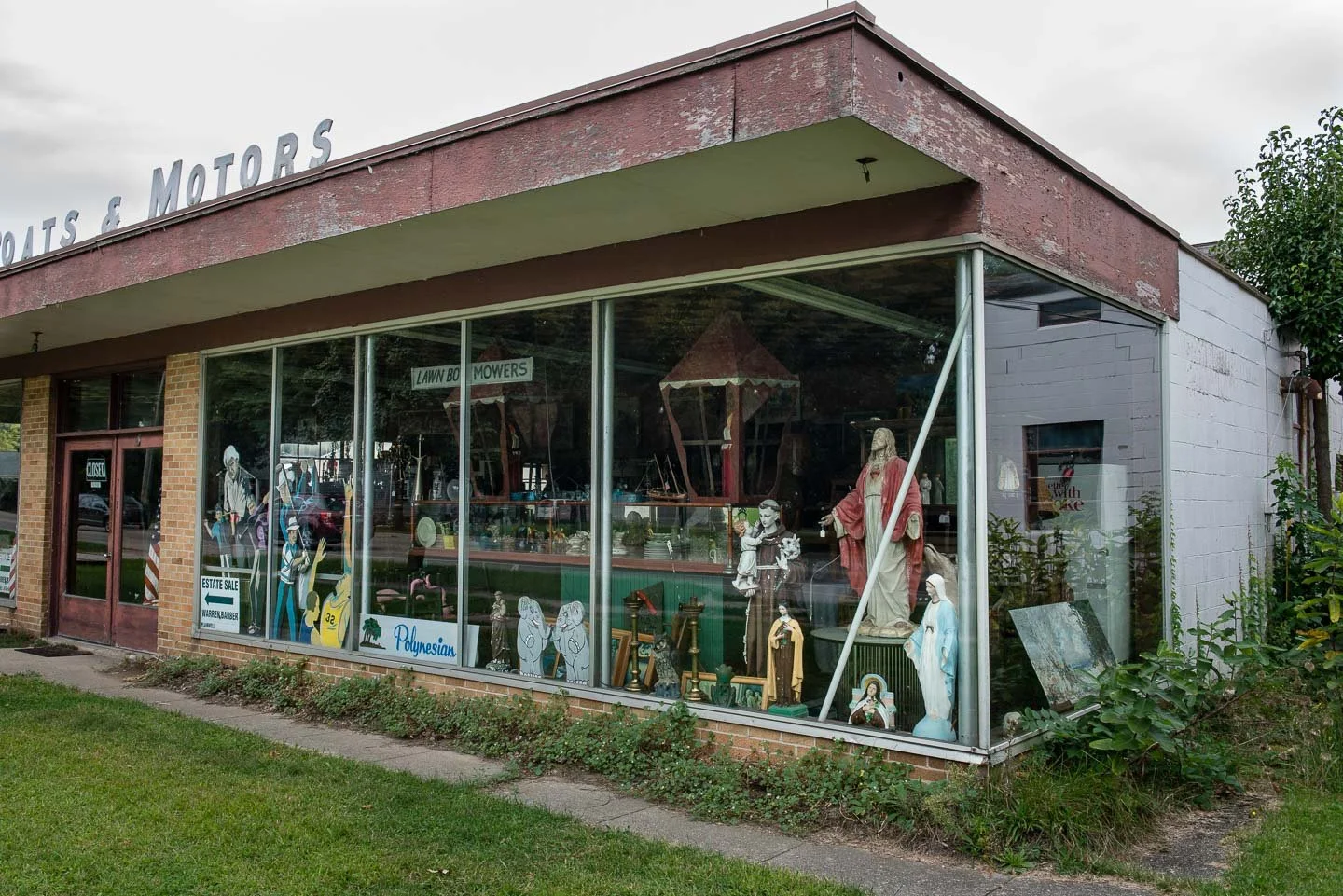 Antique store window display with religious statues, figurines, and vintage items inside. The store has a large glass front with signs for estate sale and Polynesian merchandise. Exterior shows brick and concrete walls with greenery nearby.