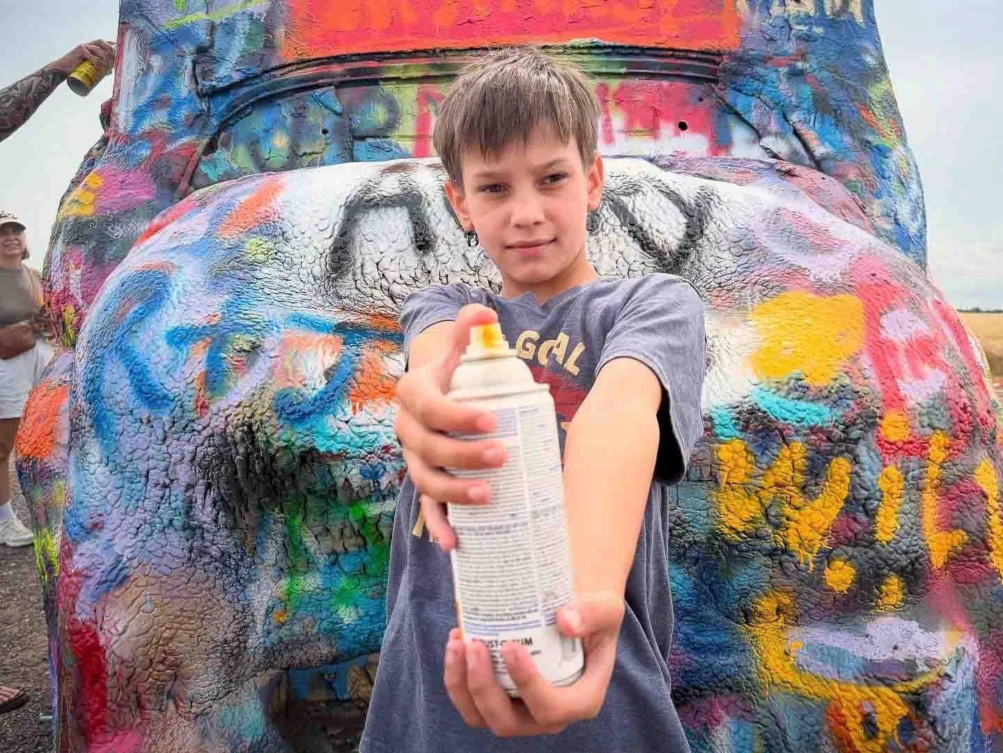 Young boy holding spray paint can standing in front of a colorful, graffiti-covered car hood, with a woman in the background smiling.