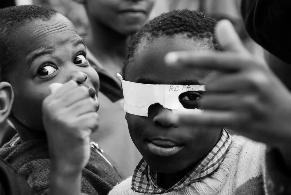Two boys flash peace signs and pose for a photo, one wearing a paper blindfold with writing and the other with wide eyes, on a group of children.