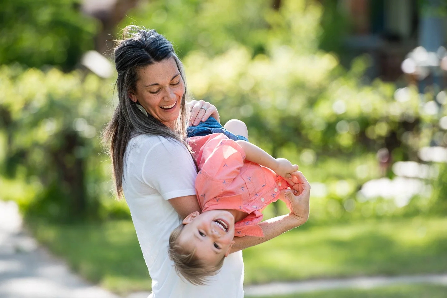 A woman smiling and playing with her young daughter outdoors in a park, the woman holding the girl upside down.