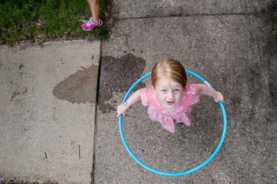 A young girl with red hair standing inside a blue hula hoop on a concrete surface, looking up at the camera with a confused expression. Part of another child's legs and purple shoes are visible in the top left corner.