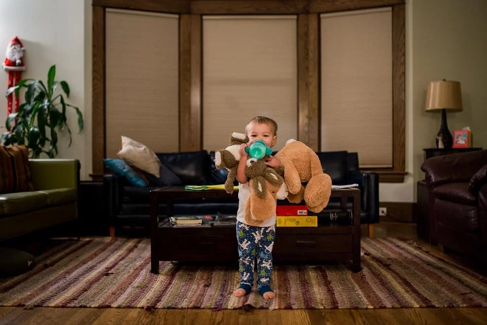 A young child standing in a living room, holding a stuffed monkey and a teddy bear, wearing pajamas with constellations, surrounded by furniture and Christmas decorations.