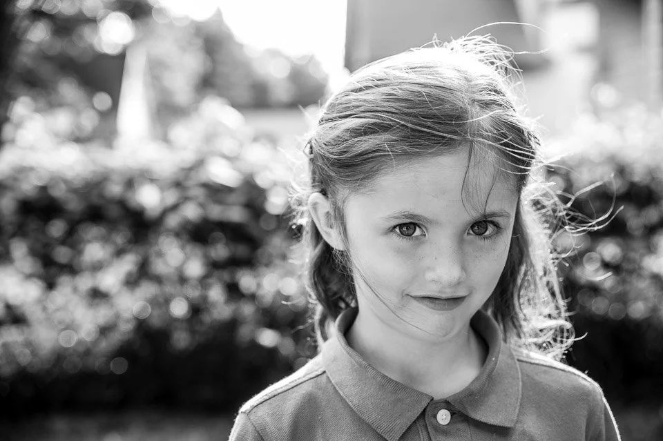 A young girl with light hair and a slight smile, standing outdoors in a garden or park, captured in black and white.