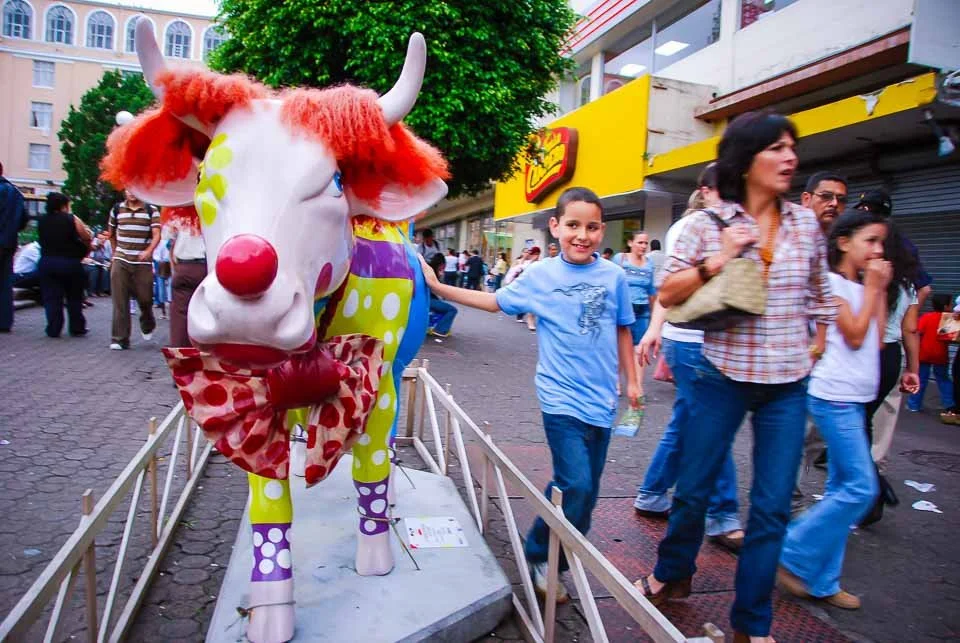 A person in a cow costume with a colorful, polka-dotted design, red hair, a large red nose, and a red bowtie, walking on a city street with pedestrians, storefronts, and green trees in the background.