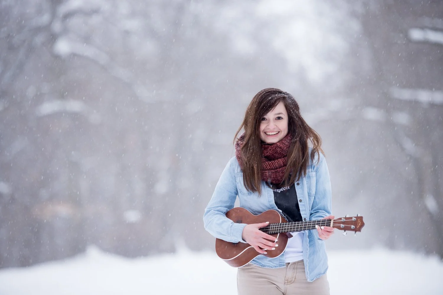 Young woman playing ukulele outdoors in snowy landscape, smiling with brown hair and wearing a denim jacket and red scarf.
