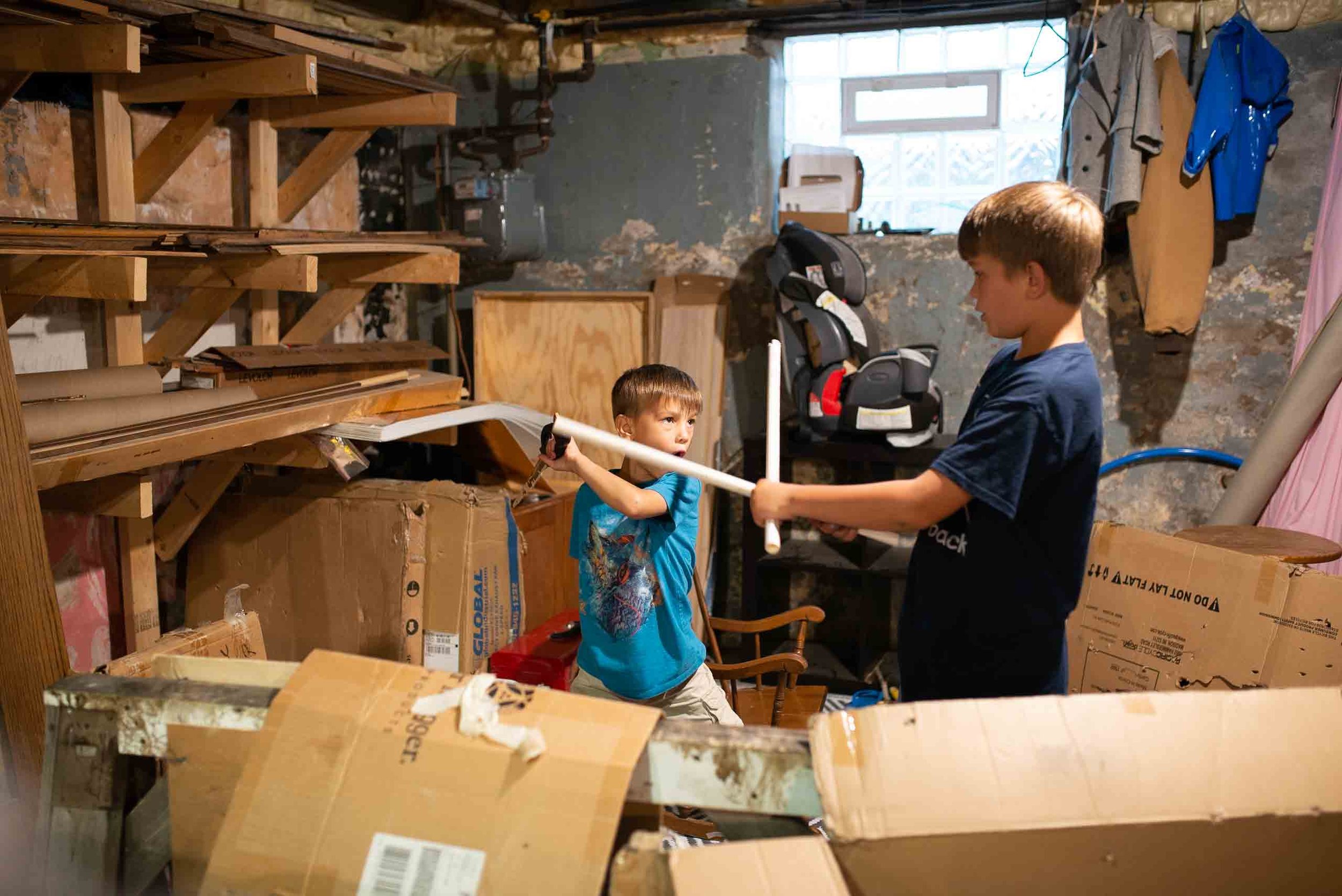 Two boys, one younger and one older, are playing with a plastic toy sword in a cluttered garage. The younger boy is standing on a chair, holding the sword while the older boy stands across from him. The garage has boxes, wooden panels, and a high cha