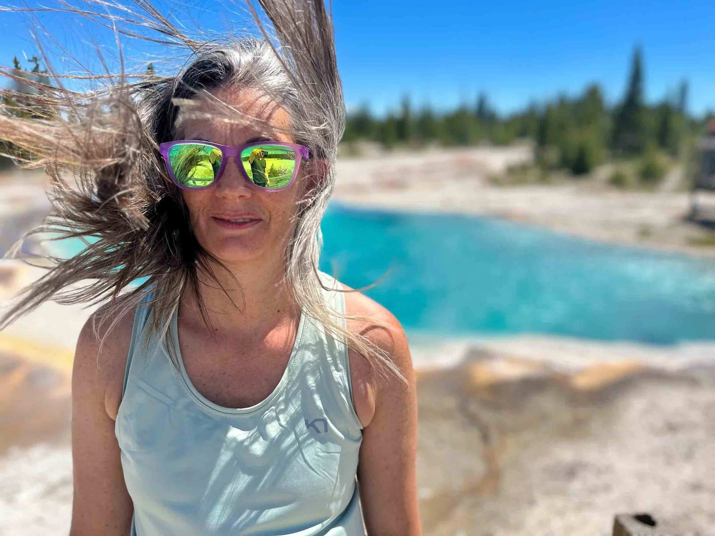 Woman with windblown hair wearing reflective sunglasses and a light-colored tank top at a beach or lakeside.
