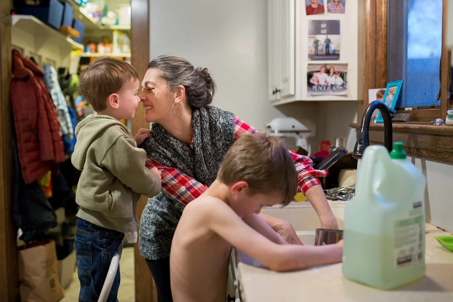 A grandmother with dark hair in a ponytail and hoop earrings, smiling as she leans close to a young boy with short brown hair, in a kitchen. Another shirtless boy is washing dishes at the sink. The background shows kitchen cabinets, photos on the wal