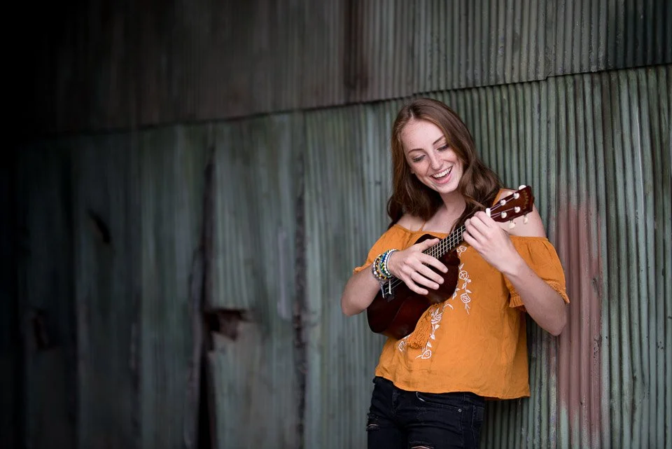 A young woman with long brown hair smiling and playing a ukulele against a wooden fence.