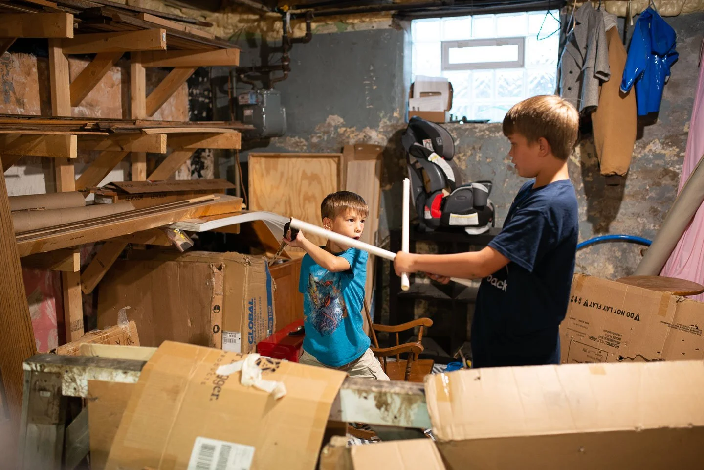 Two boys play with a toy sword and shield in a cluttered basement or storage room surrounded by boxes, wood, and household items.