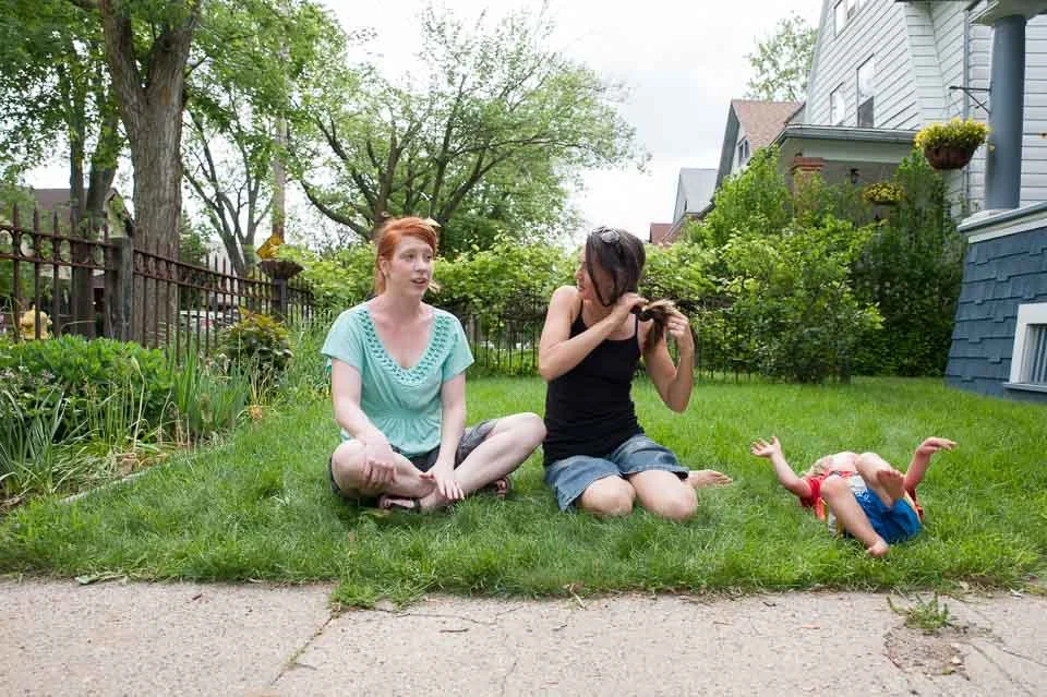 Two women sitting on the grass in a backyard, one with red hair wearing a teal top, and the other with dark hair wearing a black top, with a baby lying on the grass nearby.