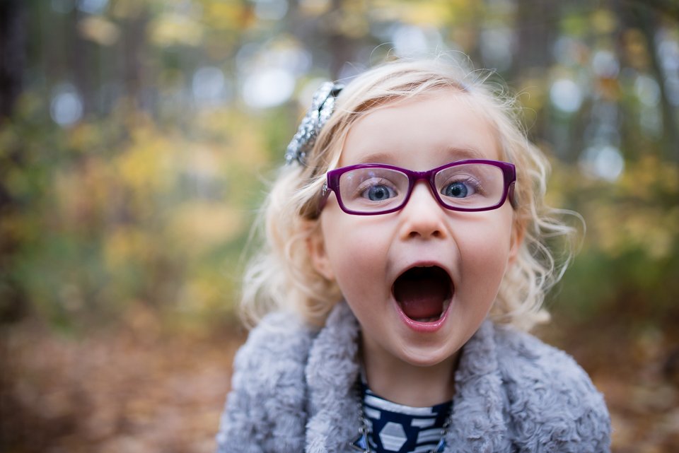 A young girl with glasses and curly blonde hair in a forest with autumn leaves, looking surprised or excited.