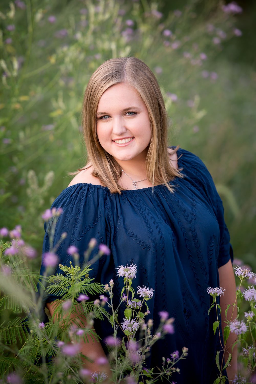 A young woman with shoulder-length blonde hair and blue eyes smiling while standing in a field of purple and white flowers, wearing a navy blue off-the-shoulder top.