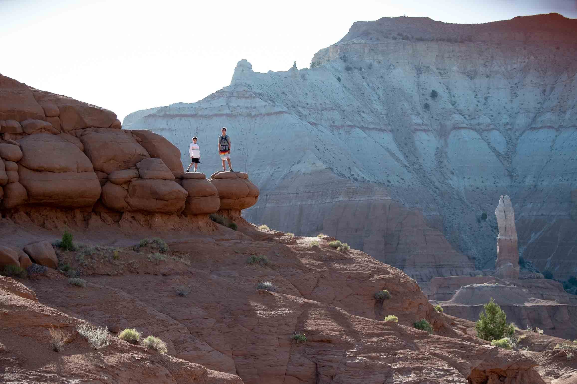 Two hikers standing on a rock ledge in a desert canyon with layered colorful cliffs in the background.