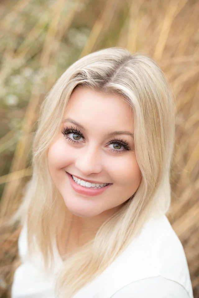 Close-up of a smiling young woman with blonde hair and blue eyes outdoors with tall grass in the background.