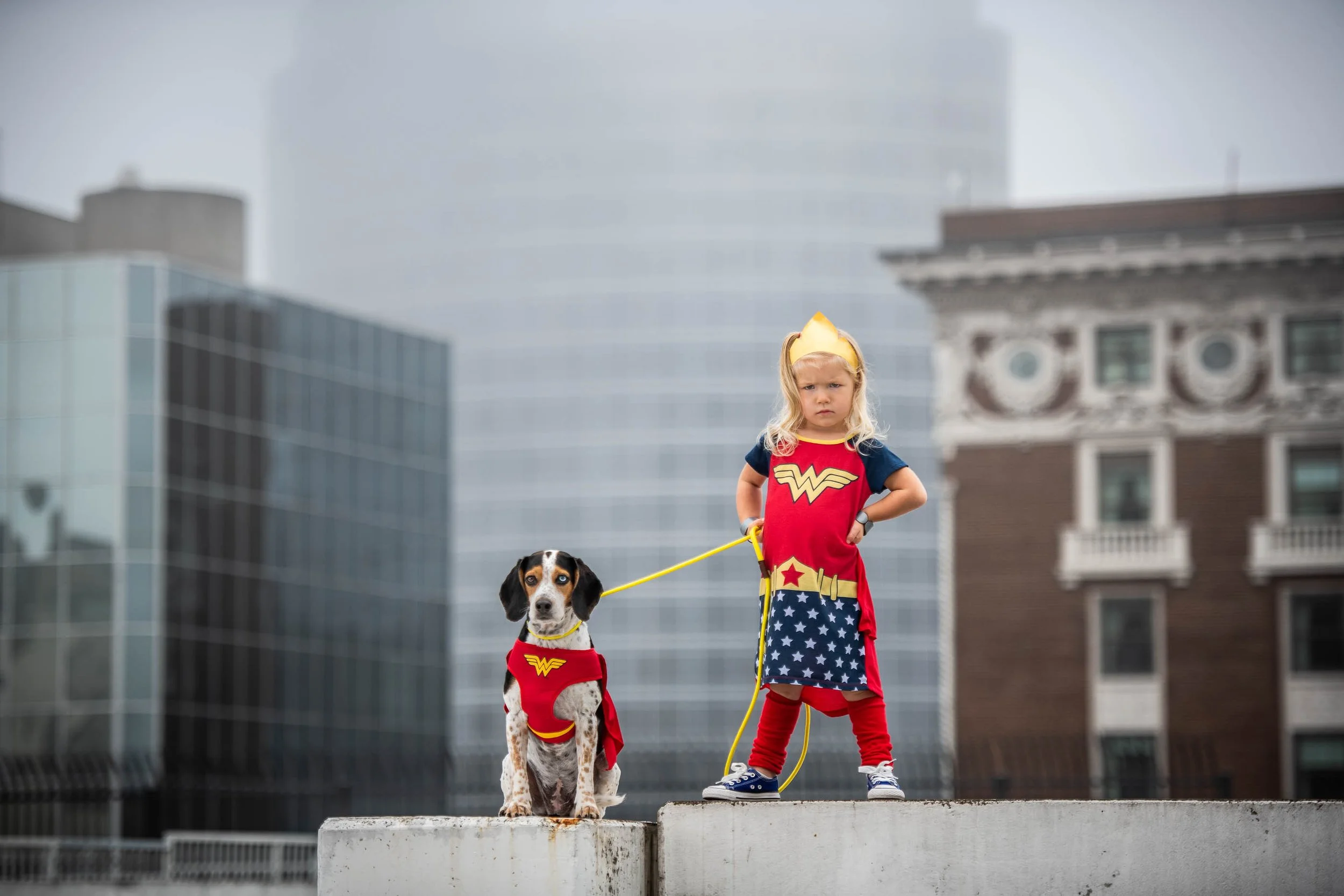 Young girl dressed as Wonder Woman standing with one hand on her hip and holding a leash attached to a dog also dressed as Wonder Woman, on a rooftop with city buildings in the background.