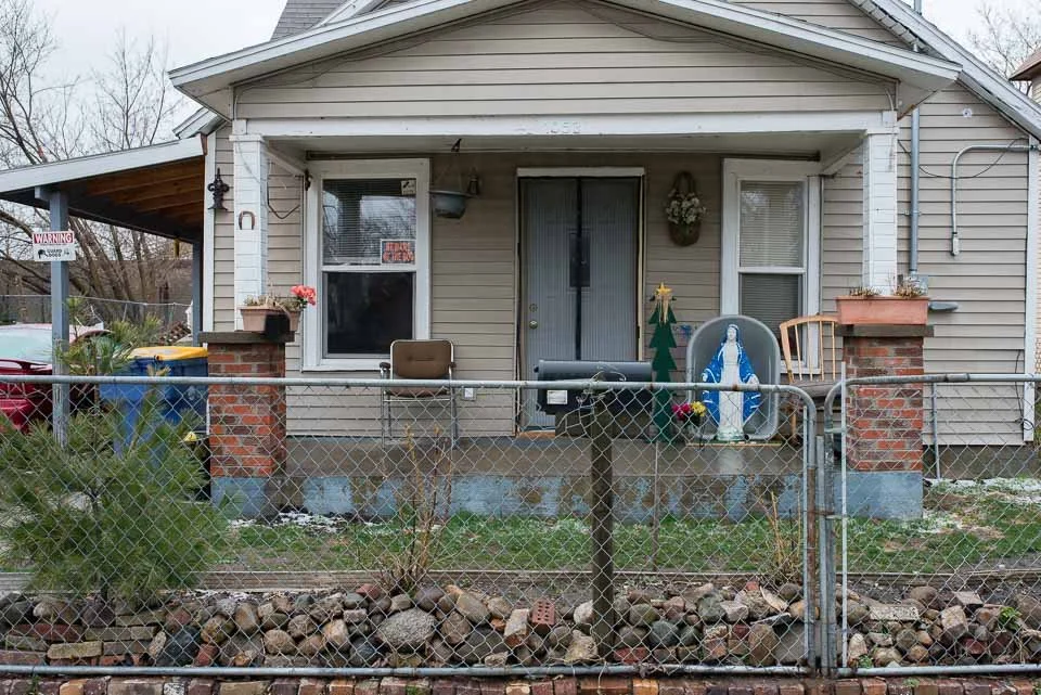 Front porch of a house with a chain-link fence, potted plants, a cardboard box, and religious decorations, including a statue of the Virgin Mary.