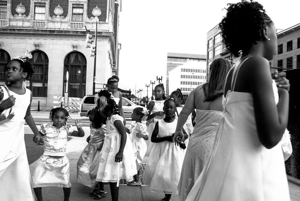 Group of young girls and women dressed in fancy white dresses, standing and walking on city street, some looking at the camera and others looking away, in an urban area with tall buildings and traffic lights.
