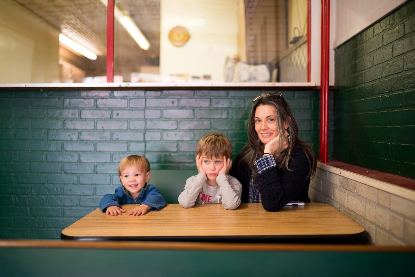 A woman with two young boys sitting at a wooden table in a restaurant, with a brick wall and a window behind them.