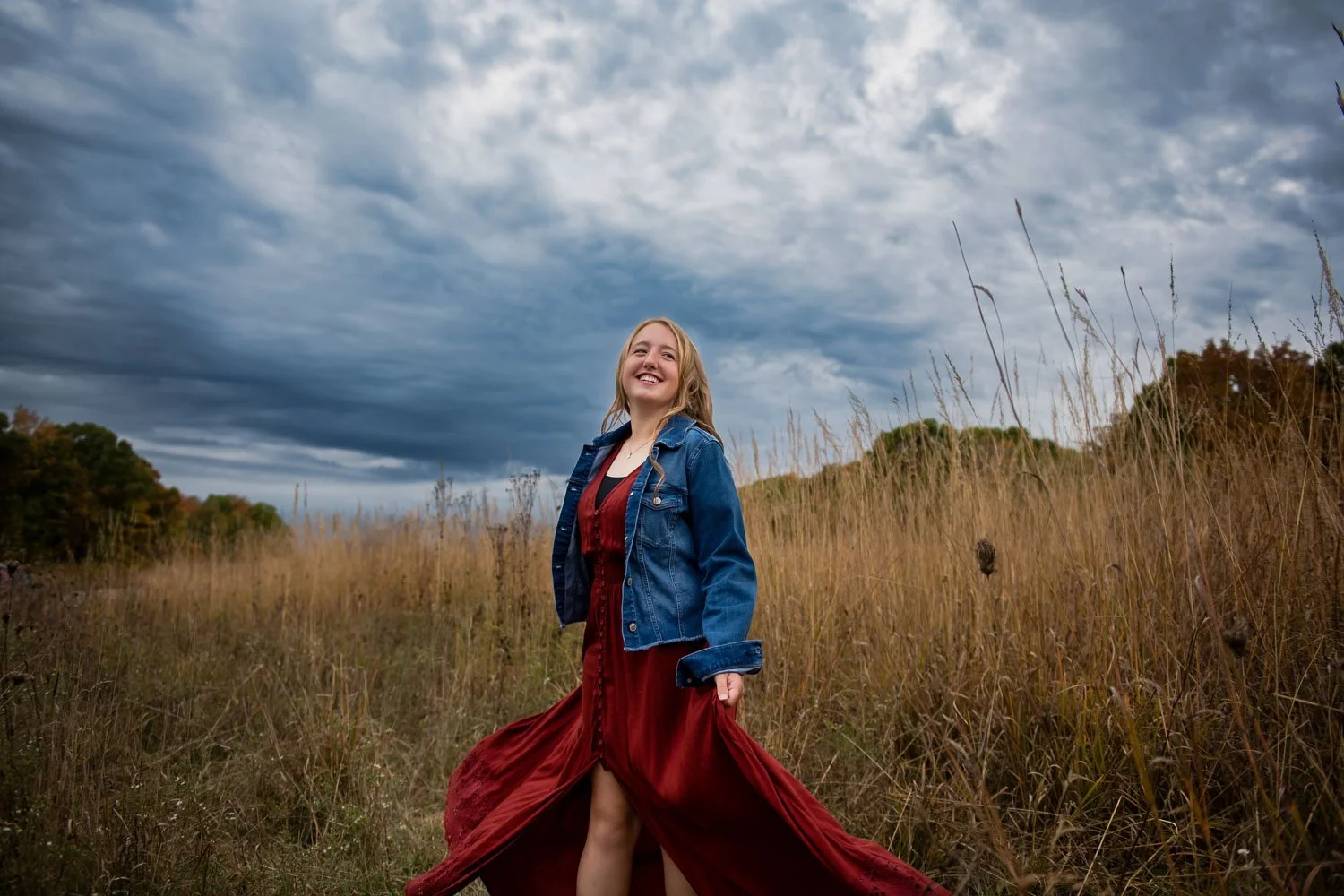 A young woman in a red dress and denim jacket standing in tall grass outdoors under a cloudy sky, smiling.